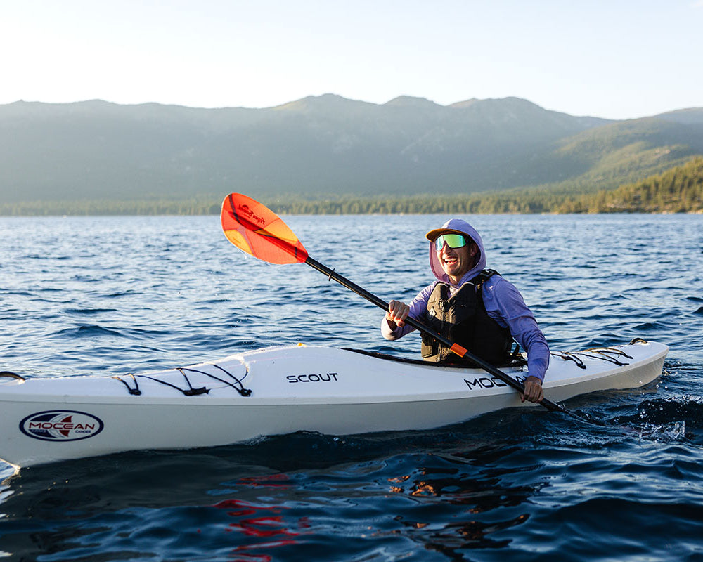 A person paddling in a white Mocean Scout kayak on a blue lake with mountains in the background, holding an orange paddle.